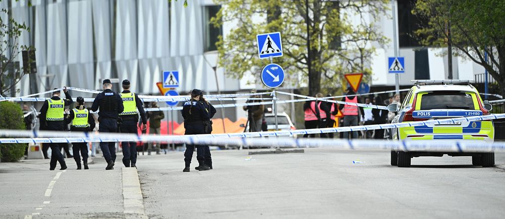 Poliser och avspärrningar vid en gata, polisbil till höger och byggnad i bakgrunden.
