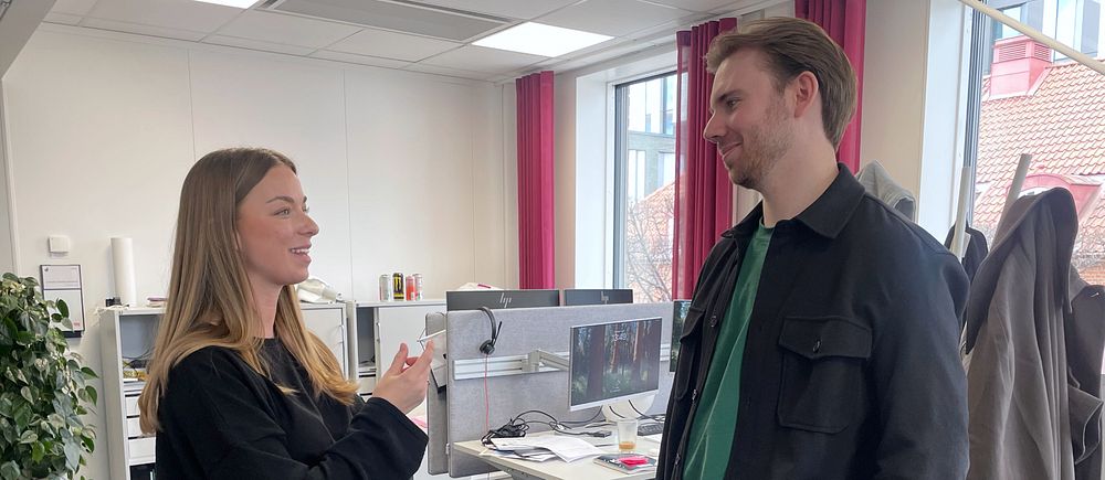 Två personer samtalar i ett kontorslandskap med stora fönster och arbetsplatser i bakgrunden.