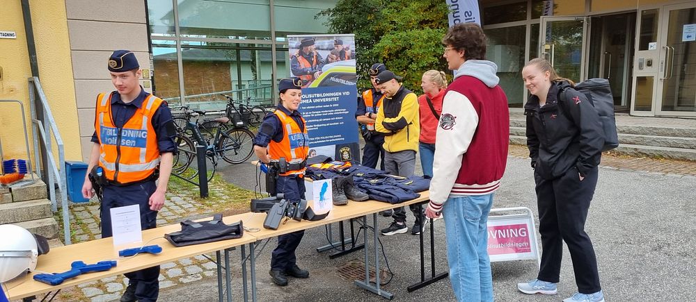 Två polisstudenter står bakom ett bord och en man med röd jacka står framför