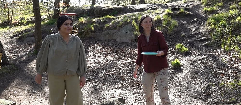 Två unga kvinnor står och spelar discgolf i en solig skogsmiljö i Slottsskogen.