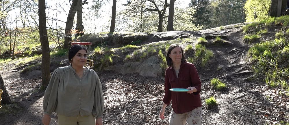 Två unga kvinnor står och spelar discgolf i en solig skogsmiljö i Slottsskogen.