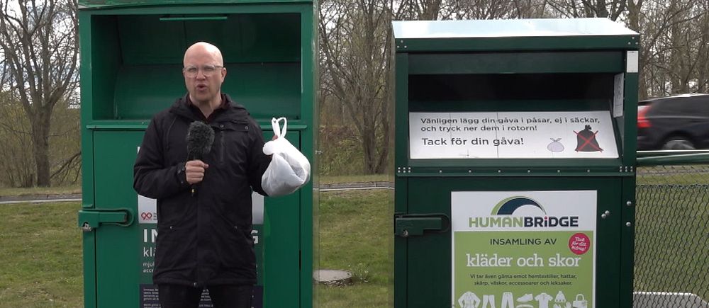 En reporter håller upp en plastpåse med kläder i. Han står framför gröna insamlingsboxar.