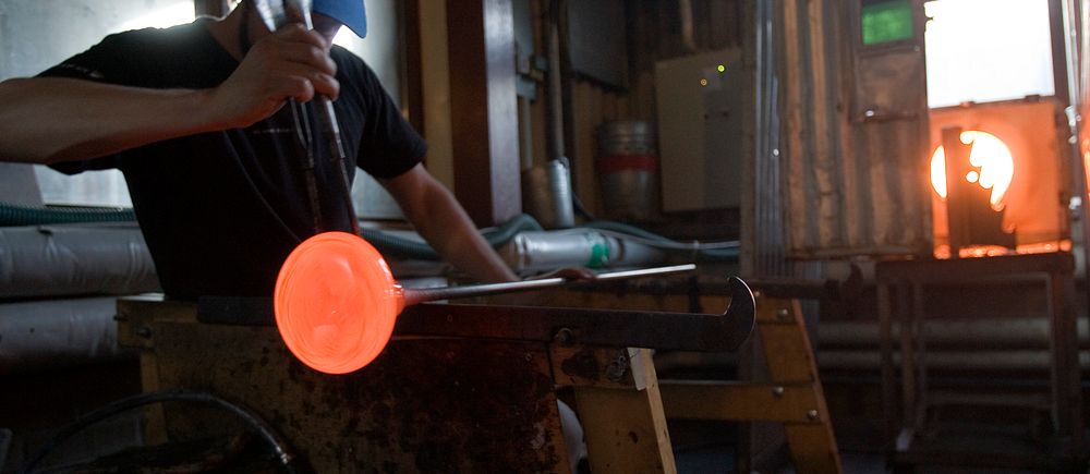 Glasblåsare formar glödande glas i en verkstad med ugn i bakgrunden.