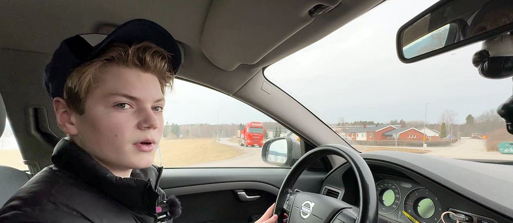 Ung kille kör bil, utomhusmiljö med lantliga byggnader i bakgrunden.