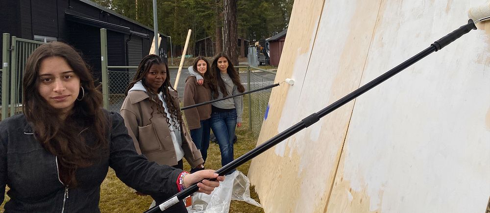Fyra personer målar en stor vägg utomhus med hjälp av färgrollers.