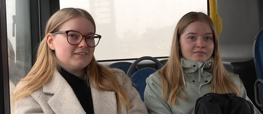 Två tjejer med långt hår sitter bredvid varann på en buss.