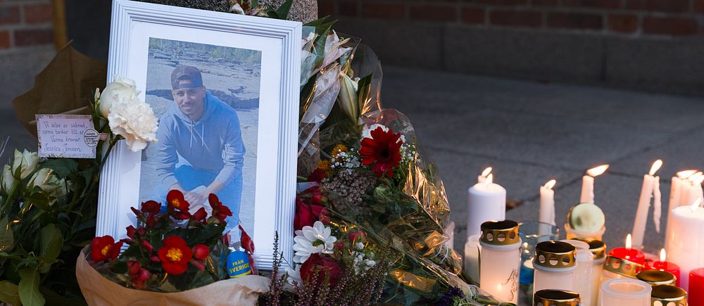 Inramad bild på 23-årig pojke, omringas av blommor och ljus på minnesplats.