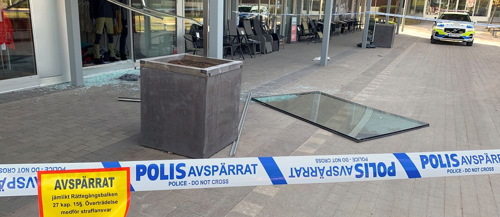 Avspärrningsband framför butik, krossat glas ligger på marken och en polisbil står parkerad i bakgrunden.