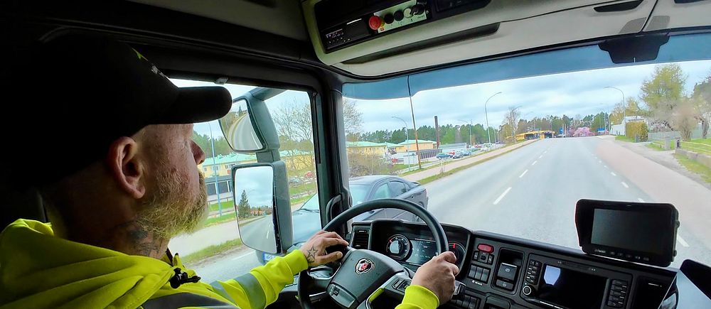 Person kör lastbil på landsväg, interiörvy av hytten.