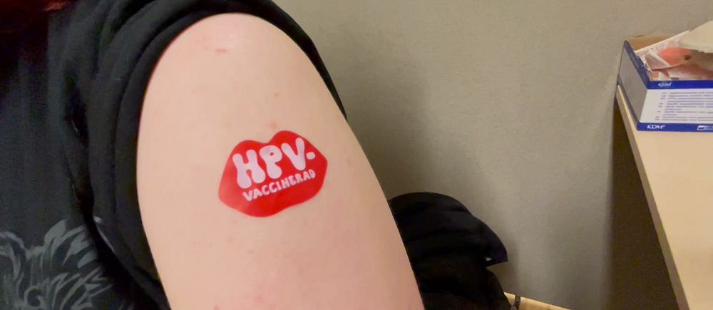 Överarm med klistermärke efter HPV-vaccination. Del av väntrum syns i bakgrunden.