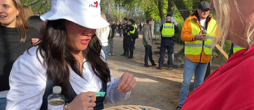 En person delar ut godis och kondomer ur korg utomhus, poliser och funktionärer syns i bakgrunden.