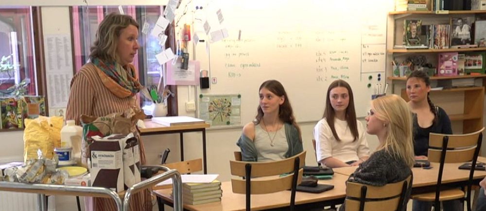 Lärare håller lektion för en grupp elever i klassrum. Elever sitter vid sina bänkar.