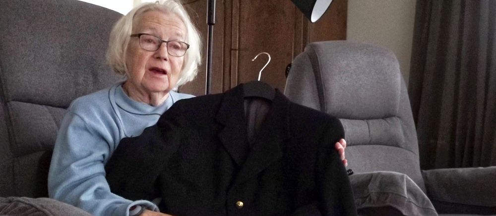 Äldre person i fåtölj håller upp en mörk kavaj med guldknappar.
