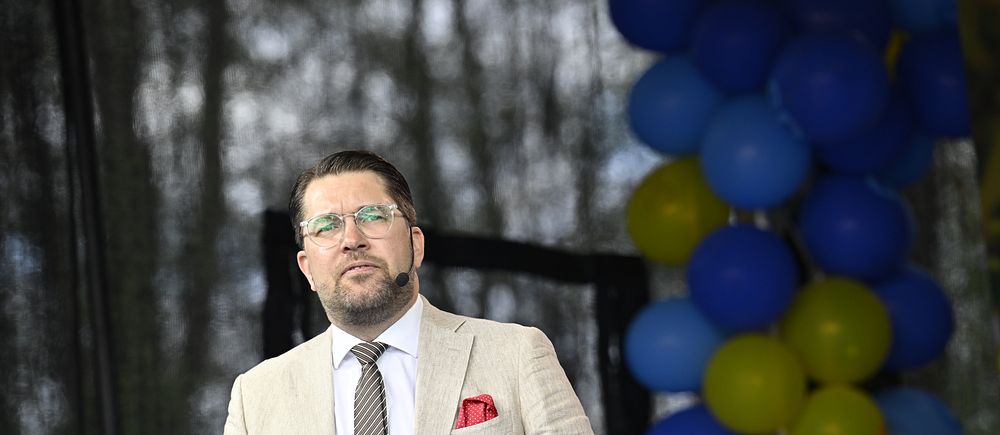 Man i beige kavaj talar på scen bredvid blå och gula ballonger.