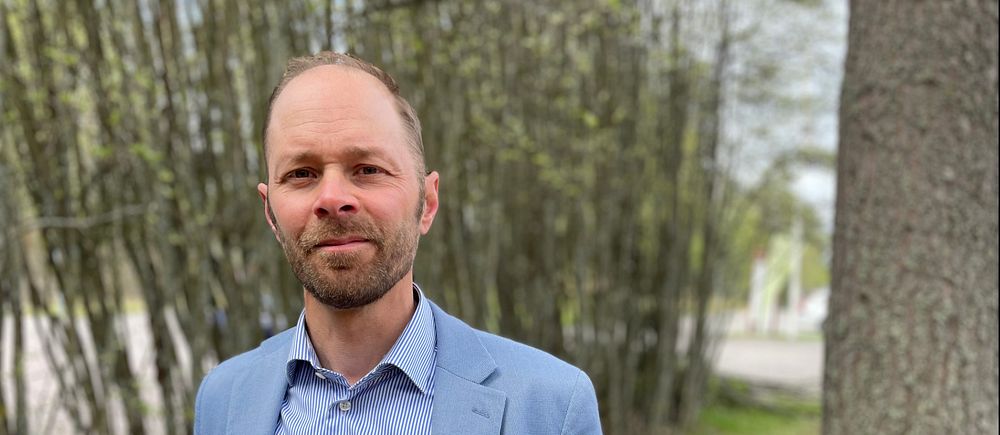 Man i ljusblå kavaj står utomhus vid träd och buskar.