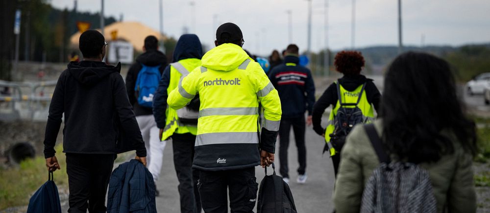 Flera Northvoltarbetare med väskor promenerar längs en väg, några bär reflexjackor där det står Northvolt på ryggen.