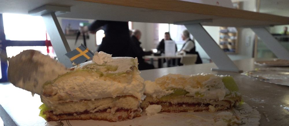 Prinsesstårta med svensk flagga på bord, suddiga människor i bakgrunden.