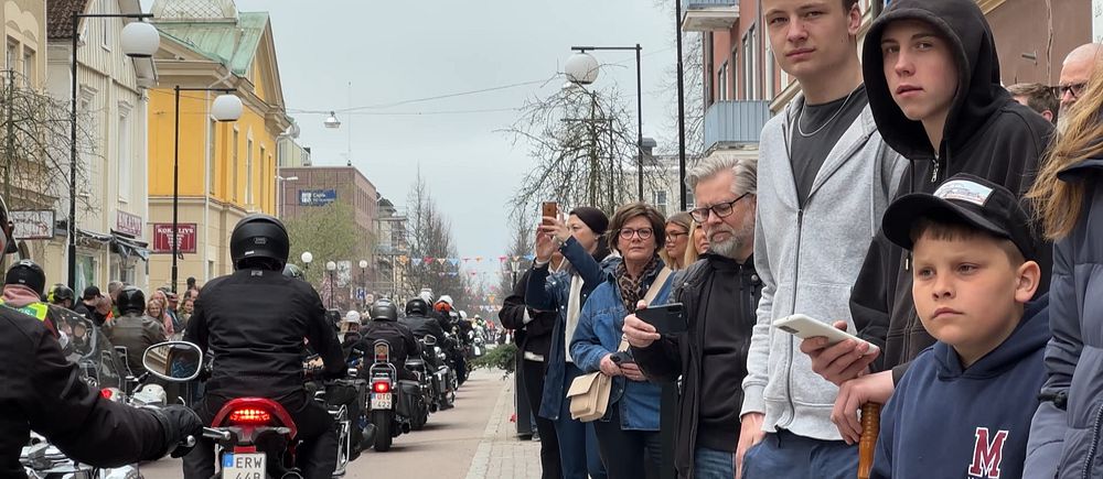 Gatumiljö med motorcyklar som kör förbi åskådare. Vit byggnad med klocktorn i bakgrunden.