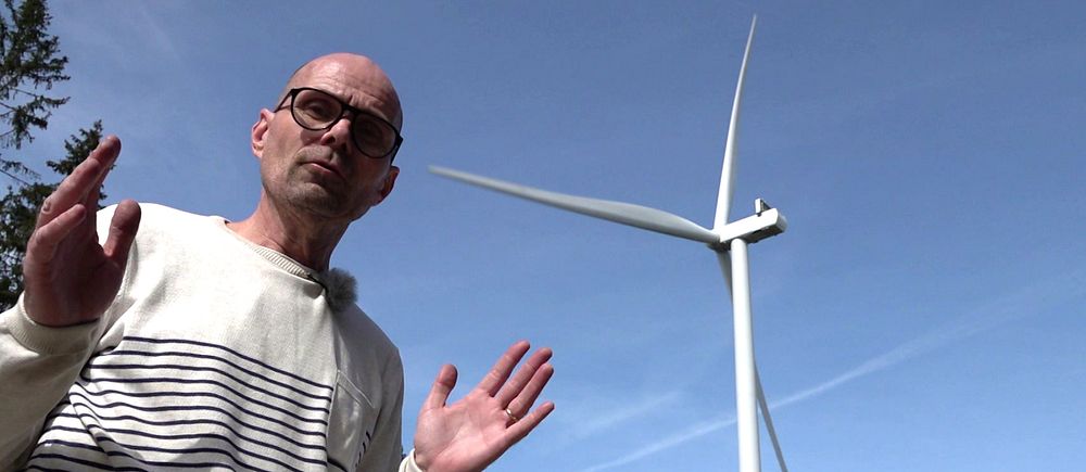 Person i randig tröja framför vindkraftverk mot blå himmel och skogskant.