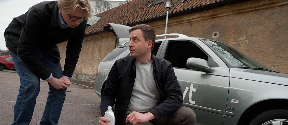 Man häller vätska ur en flaska intill bil med logga. En annan man tittar nyfiket.