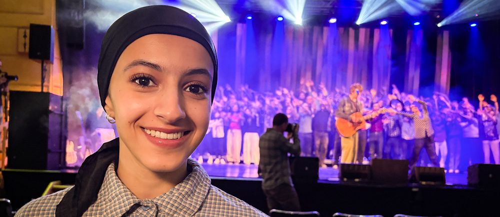 Person i förgrunden, scen med kör och musiker framför färgglada lampor i bakgrunden.