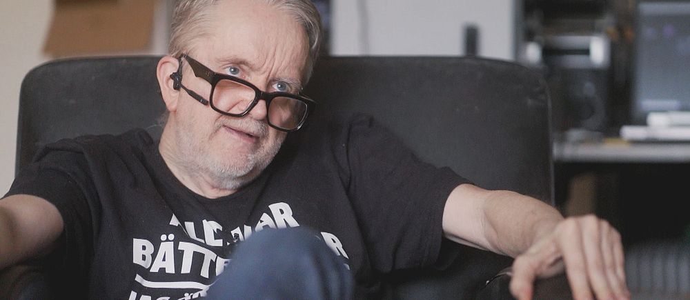 Person sitter avslappnat i en fåtölj med svart t-shirt och jeans i hemmiljö.