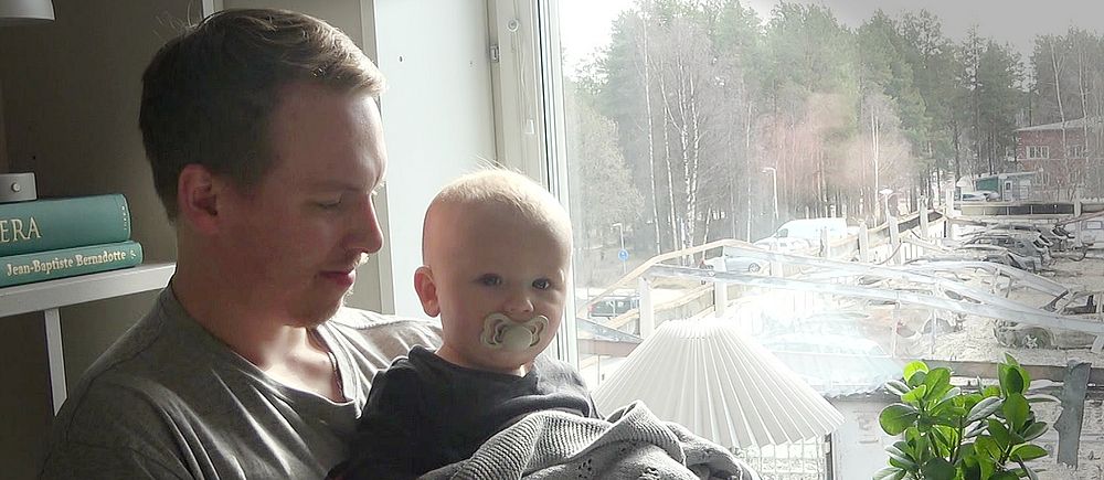 Jesper Juntto sitter hemma vid fönstret med sin son i famnen. Ute syns flera bilvrak efter den omfattande branden på Kallkällan i Luleå.