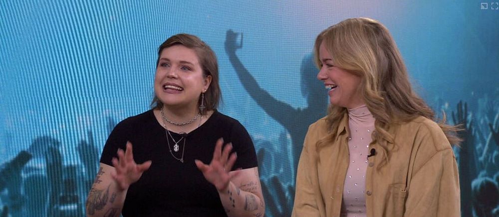 Två personer sitter vid ett bord i en tv-studio, bakom dem skymtar en konsertpublik.