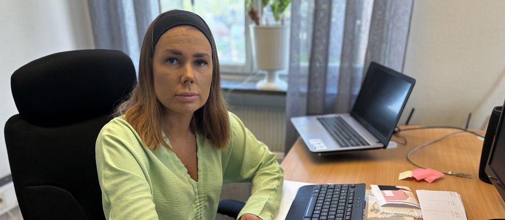 Kvinna i ljusgrön skjorta sitter vid ett skrivbord med dator och anteckningar.