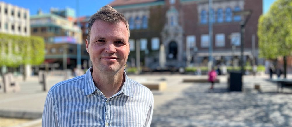 Person i randig skjorta står på soligt torg, byggnad syns i bakgrunden.