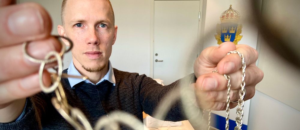 Civil polis håller upp stöldgods – kedjor och nycklar i ett kontorsrum med polissymbol i bakgrunden.
