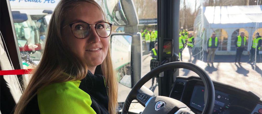Person i eldriven lastbil vid ratten, utanför står personer i gula västar vid ett tält.