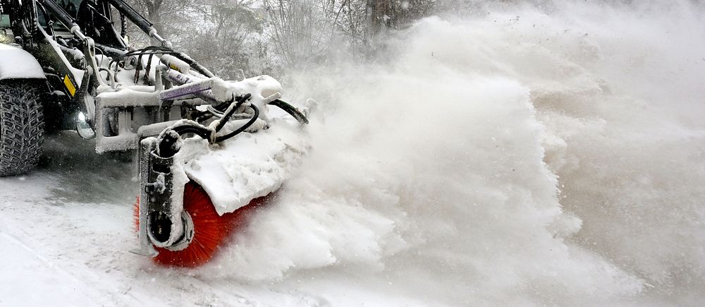Snöplog röjer snön från väg omgiven av träd i vintrigt landskap.