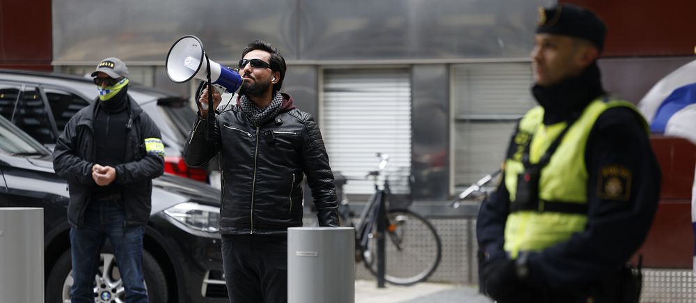 Salwan momika talar utomhus, polisen syns i förgrunden.