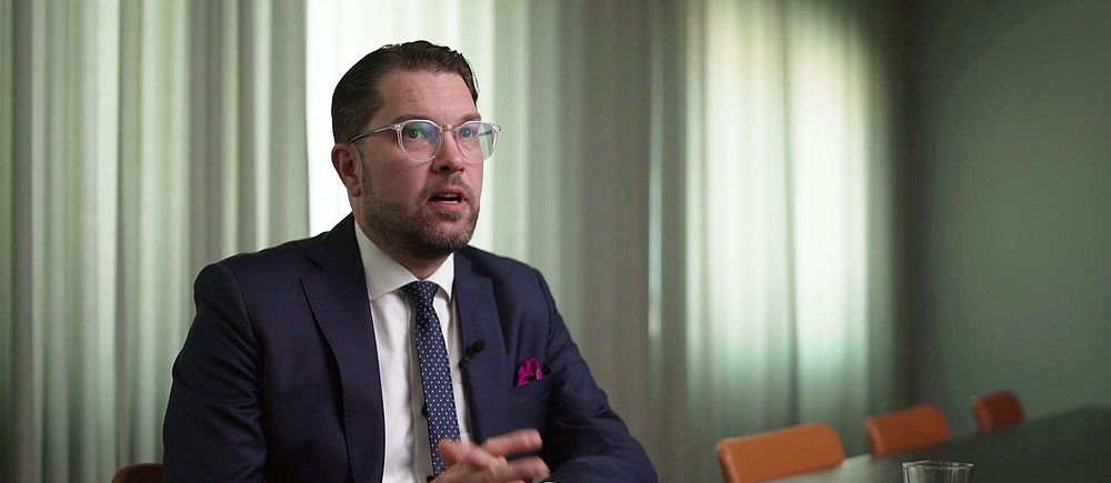 Synkbild på Jimmie Åkesson i intervju med SVT. Åkesson är klädd i kostym och sitter med händerna knäppta vid ett stort bord i ett konferensrum med fördragna garginer i dovt ljus.