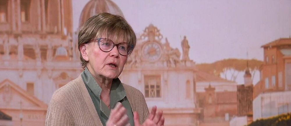 Person gestikulerar framför en bakgrund med klassisk arkitektur och kupolprydda byggnader. I bild syns Vatikankännaren Ulla Gudmundsson.