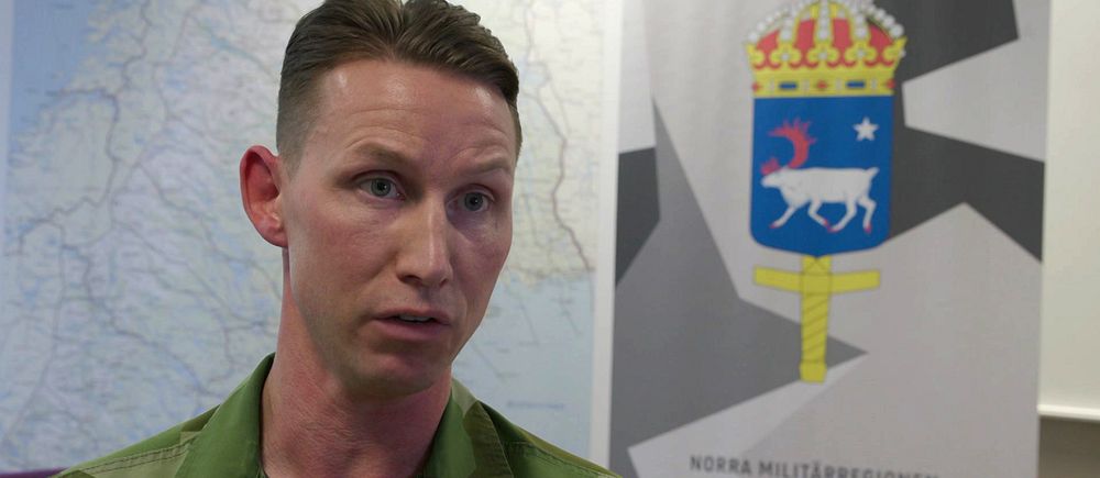 Person i militär uniform framför en kartbild och en skylt med Försvarsmaktens emblem.