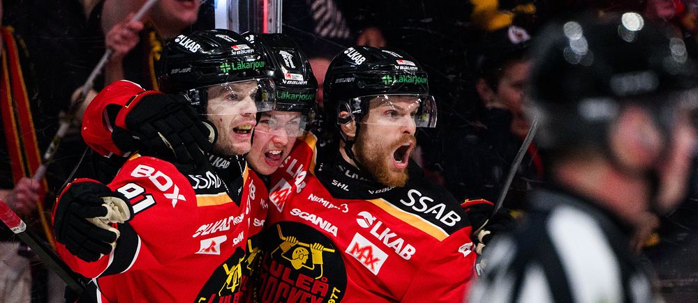 Två ishockeyspelare i röda tröjor firar ett mål, domare syns i förgrunden.