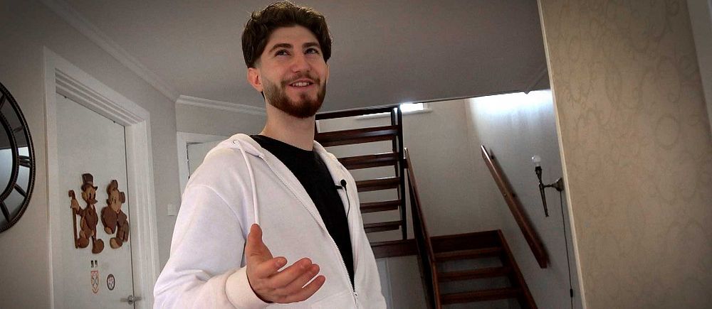 Alexander Bisso i vit hoodie står i hallen framför trappa, räcker ut handen mot kameran.