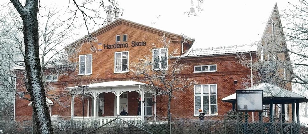 Röd tegelbyggnad som utgör Hardemo Skola, omgiven av snötäckta träd och en vinterlandskap. Byggnaden har en traditionell stil med en veranda och flera fönster. En del av skolan är annoterad med skolans namn.