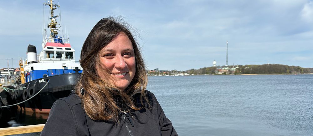 Elena Peverada står i svart jacka står vid vattnet, fartyg och kuststad syns i bakgrunden.