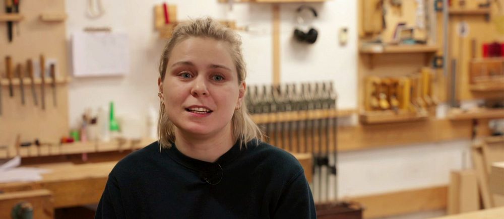tjej i mörk tröja sitter i en snickarverkstad med verktyg på väggen i bakgrunden