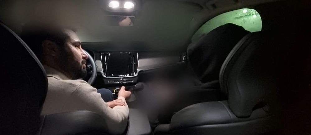 En person sitter vid ratten i en bil interiör i mörker. Till höger sitter en man med huvan uppdragen så man inte ser ansiktet.