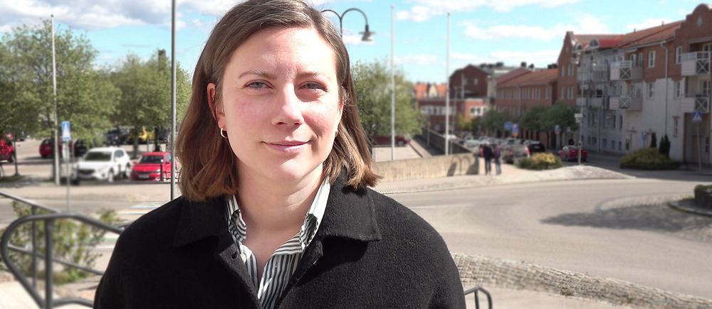 Person med axellångt hår står utomhus vid ett torg med byggnader och träd i bakgrunden.