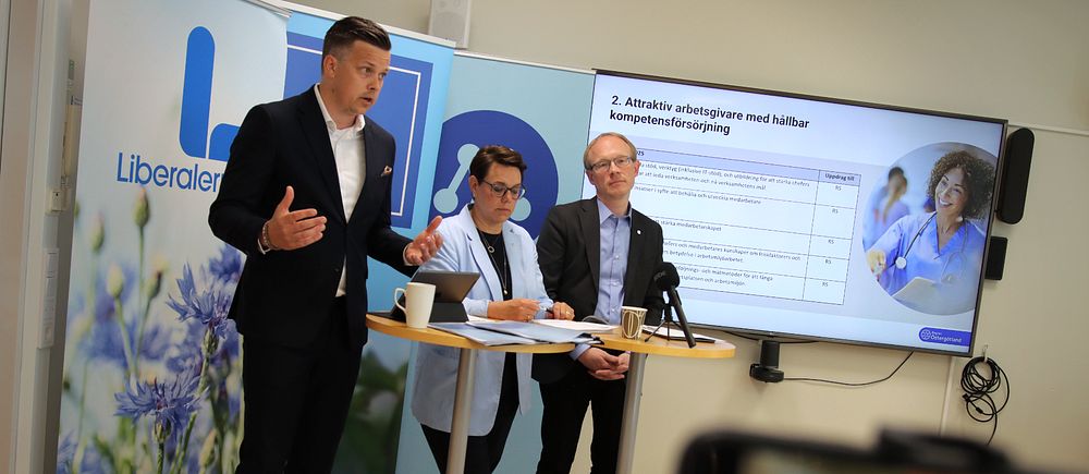 Tre politiker står framför en skärm i ett konferensrum och håller en presentation. En av de, en man i kavaj och vit skjorta gestikulerar med händerna.