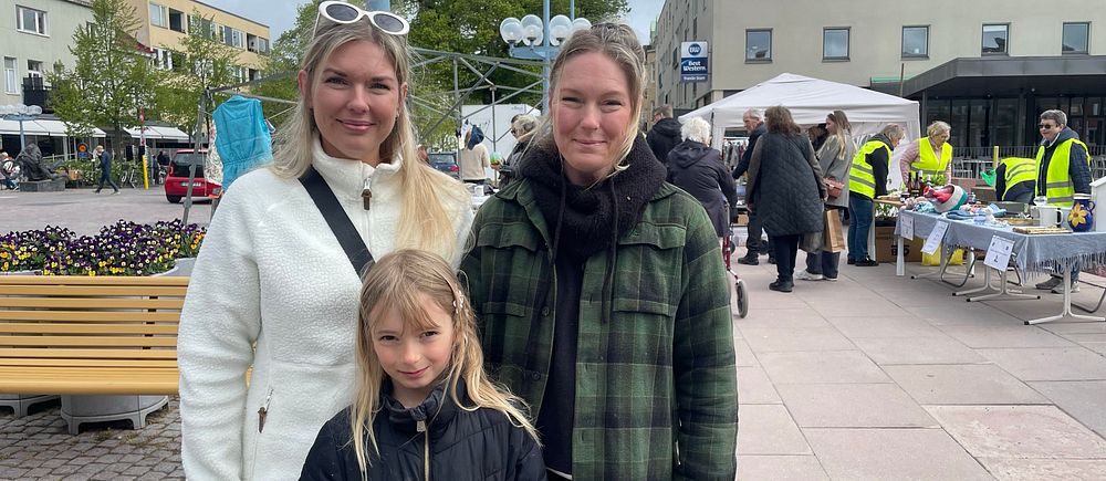 Tre personer står på ett torg, marknadsstånd och personer i västar syns i bakgrunden.