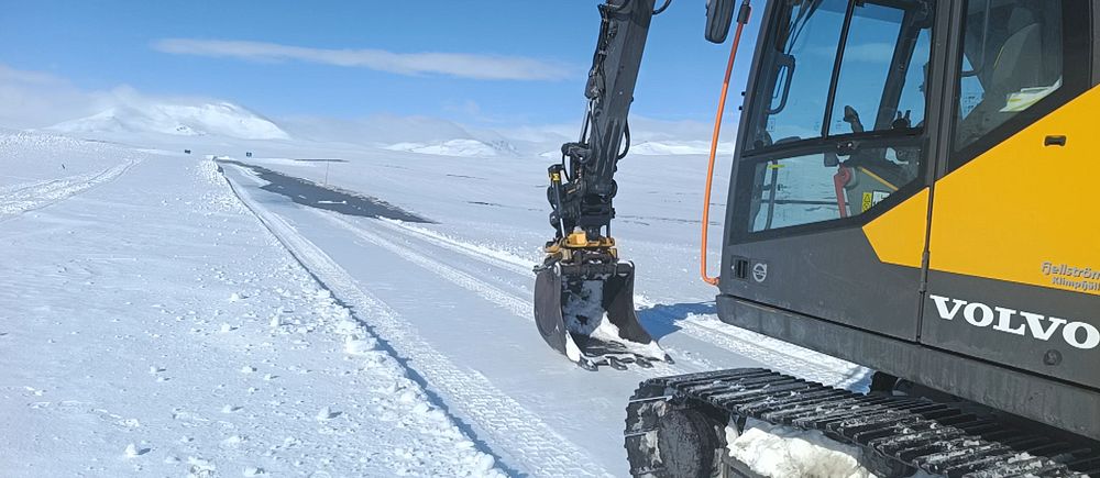 Grävmaskin från Volvo röjer väg i snötäckt landskap under klarblå himmel.