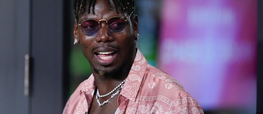 Paul Pogba klädd i en rosa skjorta med mönster och flera kedjor hänger runt halsen. Bakgrunden är suddig men visar en skarp färgkontrast som antyder en evenemangsmiljö.