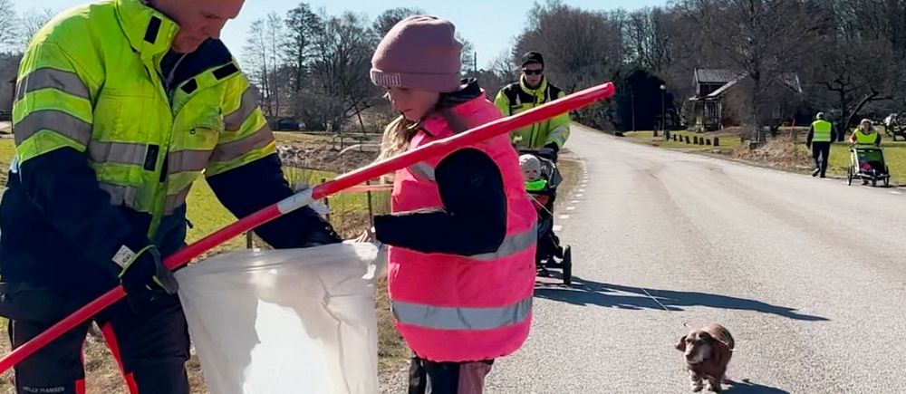 Två personer städar vägkant, en liten hund står på vägen.