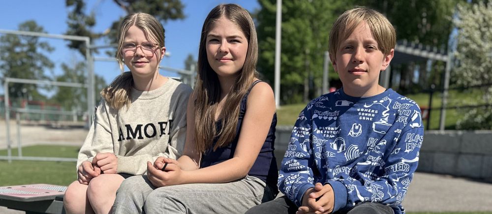 Tre elever, två tjejer och en kille som går i årskurs fem sitter på en bänk utomhus på skolgården.